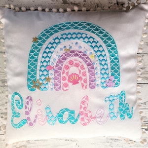 Könnte beinhalten: Ein weißes Kissen mit einem Meerjungfrauen-Regenbogen-Design. Der Regenbogen besteht aus türkisfarbenen, violetten und rosafarbenen Schuppen mit goldenen Sternen und einer Muschel. Der Name "Elizabeth" ist unter dem Regenbogen in türkisfarbener, violetter und rosafarbener Schrift gestickt.