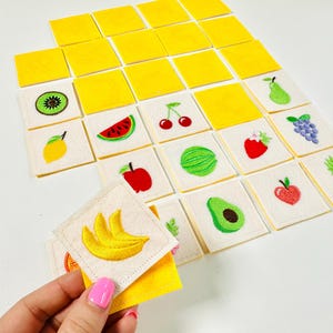 Könnte beinhalten: Eine Sammlung von bestickten Quadraten mit Obstmotiven, darunter Kiwi, Wassermelone, Kirschen und mehr. Die Quadrate sind auf einer weißen Oberfläche angeordnet, mit einer Hand, die einen Stapel Bananenquadrate hält. Die Quadrate sind gelb und weiß.