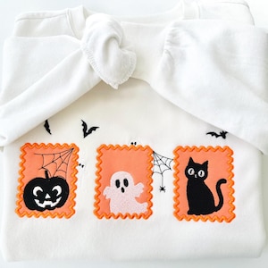May include: White sweatshirt with Halloween-themed embroidery. Features three orange postage stamp-style patches with a black jack-o'-lantern, a pink ghost, and a black cat. Black bats and spiderwebs are also embroidered.