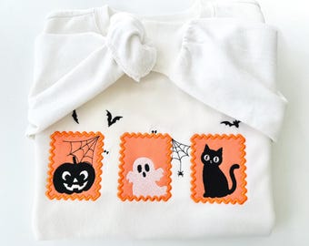 Pode incluir: Camisola branca com bordados de Halloween. Apresenta três patches laranja em forma de selo postal com uma abóbora preta, um fantasma rosa e um gato preto. Morcegos pretos e teias de aranha também são bordados.