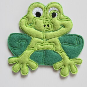 May include: A green felt frog with embroidered details, including eyes, a mouth, and a patterned body. The frog is a puzzle piece with multiple sections.