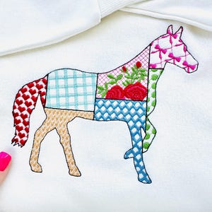 Puede incluir: Una sudadera blanca con un diseño de caballo de patchwork bordado en la parte delantera. El caballo está hecho de cuadrados de diferentes colores, incluyendo rojo, azul, verde y amarillo.