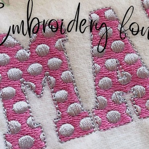 以下が含まれることがあります： ピンクと白のドット柄の刺繍された文字「M」と「A」。文字は銀色の糸で縫われています。文字は白い布地に刺繍されています。文字の上に「Embroidery font」というテキストが筆記体で書かれています。
