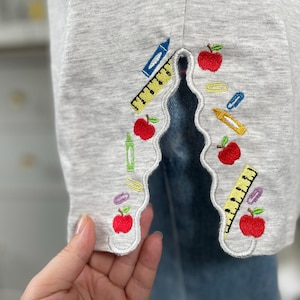 May include: Close-up of a light gray garment with a scalloped edge, embroidered with school-themed motifs. The design includes red apples, yellow rulers, blue crayons, and other colorful school supplies. The garment is likely a shirt or a similar item.