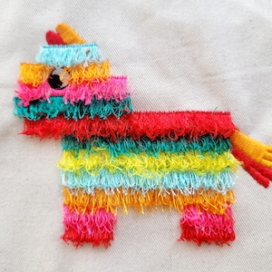 May include: A colorful embroidered pinata shaped like a donkey with a single eye. The pinata is made of red, yellow, green, blue, and orange yarn.