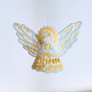 Puede incluir: Un adorno de ángel blanco y dorado con detalles de encaje. El ángel tiene alas y un halo. El adorno está colgado de una cuerda blanca.