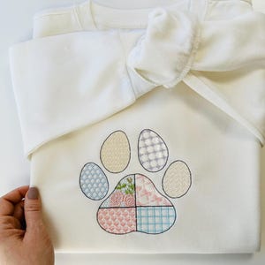 Może przedstawiać: Biała bluza z kolorowym haftowanym wzorem łapy. Łapa składa się z sześciu kwadratów w różnych kolorach z różnymi wzorami.