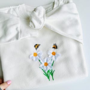 Puede incluir: Sudadera blanca con margaritas y abejas bordadas. La sudadera tiene un detalle de lazo en el cuello.