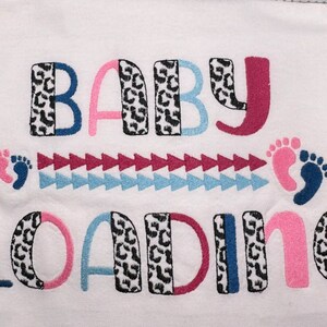 May include: White onesie with the text "BABY LOADING" in pink, blue, black, and red. The letters are decorated with a black and white leopard print. There are two footprints, one pink and one blue, above the text.