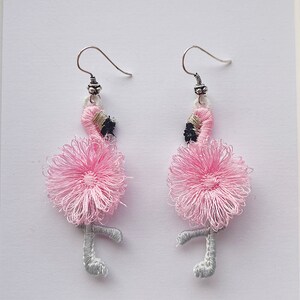 May include: A pair of pink flamingo earrings with silver hooks. The flamingos are made of pink and white thread and have black eyes.