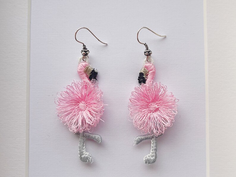 May include: A pair of pink flamingo earrings with silver hooks. The flamingos are made of pink and white thread and have black eyes.
