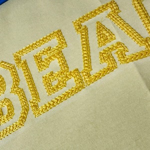 May include: Close-up of the word "BEARD" embroidered in gold thread on a beige fabric.