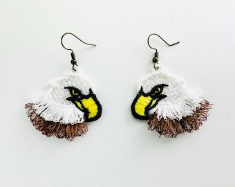 Puede incluir: Un par de pendientes hechos a mano con un diseño de cabeza de águila bordada. Los pendientes tienen una combinación de colores blanco, marrón, amarillo y negro. La cabeza del águila está detallada con un pico amarillo y un ojo negro, y los pendientes tienen un cierre de gancho.