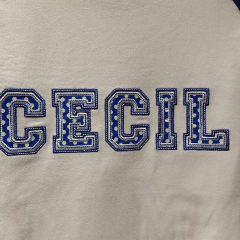 May include: White fabric with the name "CECIL" embroidered in blue and silver letters with a white polka dot pattern.