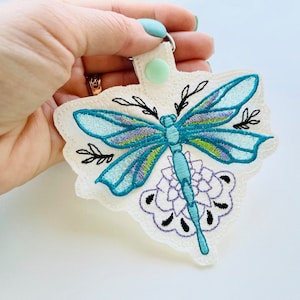 May include: A white fabric keychain with an embroidered blue dragonfly and purple flower design. The dragonfly has green accents and the flower has black outlines.