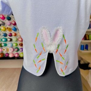 May include: A light blue t-shirt with a white bunny rabbit embroidered on the back. The bunny has pink ears and is surrounded by green and orange carrots.