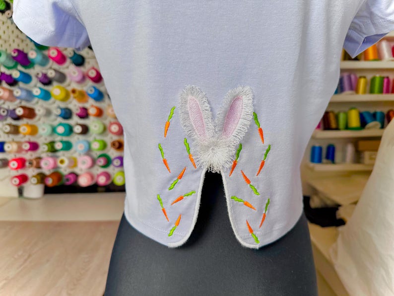 May include: A light blue t-shirt with a white bunny rabbit embroidered on the back. The bunny has pink ears and is surrounded by green and orange carrots.