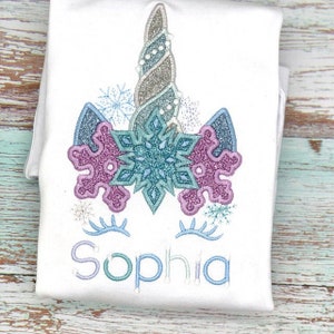 Op de afbeelding: Witte romper met een geborduurd sneeuwvlok-eenhoornontwerp. De eenhoorn heeft een zilveren glitterhoorn, blauwe en paarse sneeuwvlokaccenten en wimpers. De naam "Sophia" is onder de eenhoorn geborduurd.