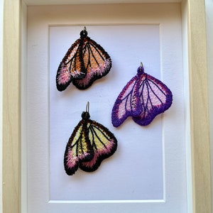 May include: Three embroidered butterfly earrings displayed in a white frame. The butterflies are pink, purple, and orange with black accents.
