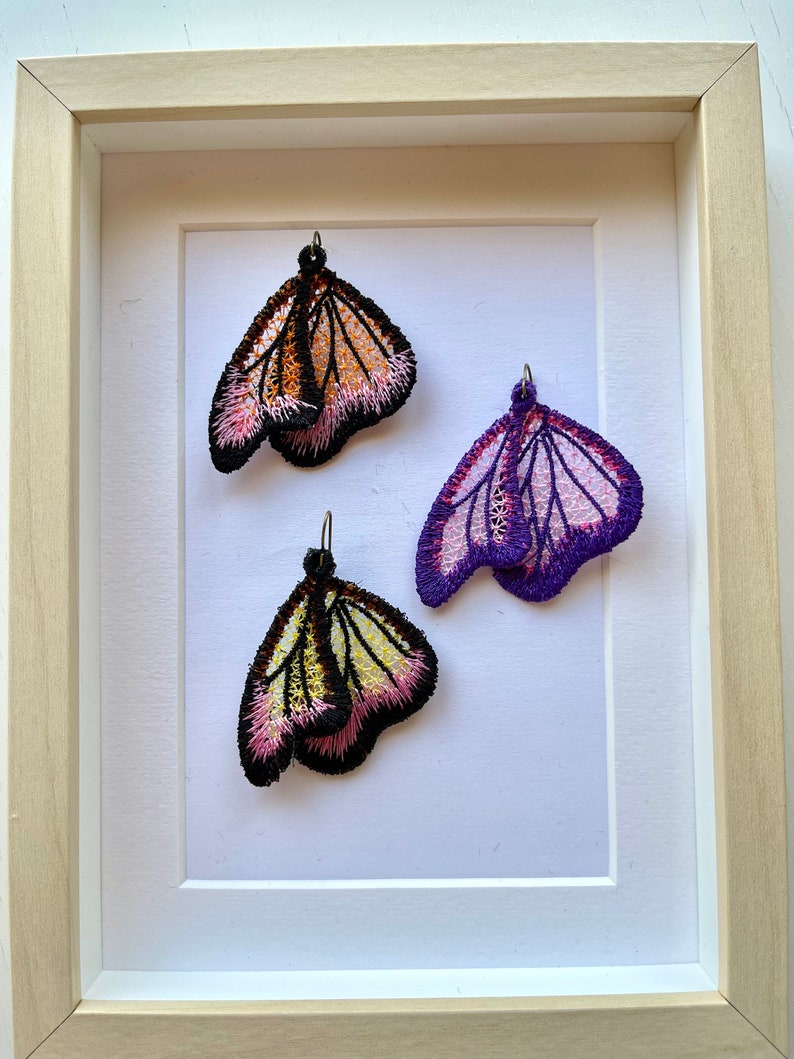 May include: Three embroidered butterfly earrings displayed in a white frame. The butterflies are pink, purple, and orange with black accents.