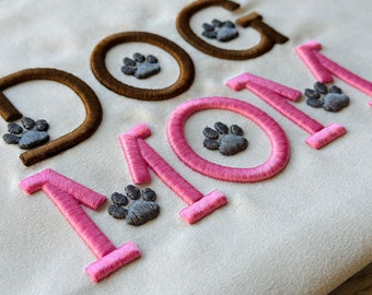 May include: Embroidered "DOG MOM" design in pink and brown with paw prints in grey.