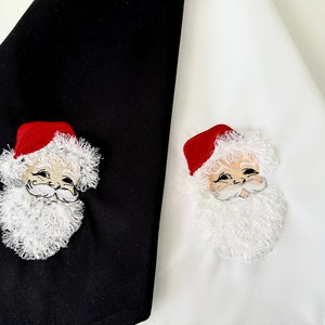 May include: Two kitchen towels with embroidered Santa Claus faces. One towel is black with a white Santa Claus face and the other is white with a white Santa Claus face.