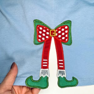 May include: A light blue shirt with an embroidered elf design. The elf has red and white striped legs, green shoes, and a red bow with white polka dots.