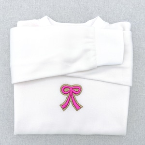 Op de afbeelding: Een gevouwen witte sweatshirt met een roze en gouden geborduurde strik op de voorkant. De lange mouwen zijn omgeslagen en de manchetten zijn zichtbaar. De sweatshirt is gemaakt van een zacht materiaal.