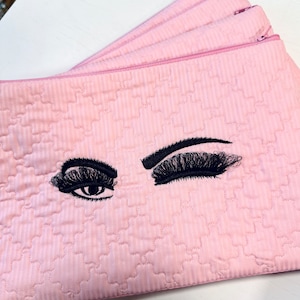 May include: Pink quilted makeup bag with embroidered eyelashes. The bag has a zipper closure.