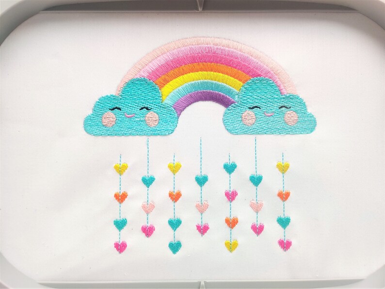 May include: Embroidered design of a rainbow with two smiling clouds and a string of colorful hearts hanging from the clouds.