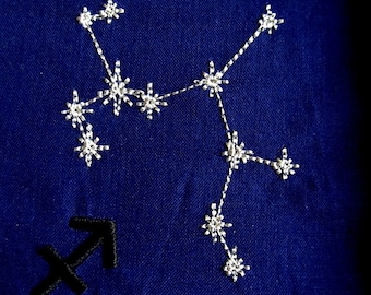 May include: A navy blue fabric with a white embroidered constellation of Sagittarius, the archer. The constellation is surrounded by a black embroidered symbol of the archer.