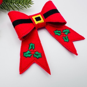 May include: A festive red bow with black accents and a gold buckle design. The bow features green embroidered holly leaves on the tails, perfect for holiday decorations. The bow is made of felt.
