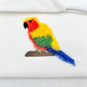 May include: Embroidered parrot in vibrant colors of red, yellow, green, and blue, perched on a brown branch. The bird's detailed design is set against a white background, showcasing the intricate stitching of the artwork.