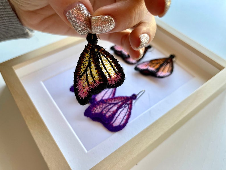 May include: Three pairs of butterfly-shaped earrings, each with a different color scheme. The earrings are made of delicate lace and feature intricate details. The colors include pink, purple, yellow, and black.