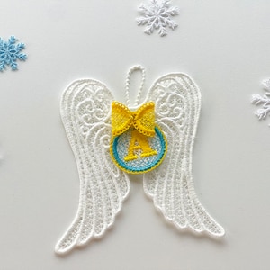 Puede incluir: Alas de ángel de encaje blanco con un lazo amarillo y un círculo azul con la letra A en el centro. Las alas están colgadas sobre un fondo blanco.