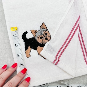 Peut inclure: Serviette blanche avec un Yorkshire Terrier brodé. Le chien est noir, beige et gris. La serviette a des rayures rouges sur le bord et mesure environ 10 cm de long.