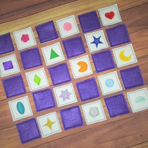 May include: A set of 20 felt squares with various embroidered shapes and symbols, including a heart, star, moon, and geometric shapes. The squares are arranged in a grid pattern on a wooden surface.