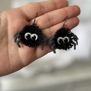 May include: A pair of black spider earrings with white eyes and grey legs. The earrings are made of a fuzzy material and have a hook closure. The spider earrings are a fun accessory for Halloween or everyday wear.