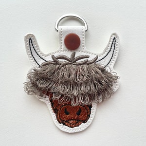 May include: White leather keychain with an embroidered brown and gray Highland cow head.