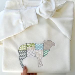 May include: A white sweatshirt with a patchwork sheep design embroidered on the front. The sheep is made up of different colored squares, including green, blue, and white.