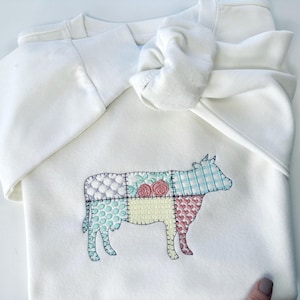 Puede incluir: Sudadera blanca con un diseño de vaca de patchwork bordado en la parte delantera. La vaca está delineada en negro y tiene una variedad de colores, incluyendo rosa, azul, verde y amarillo.
