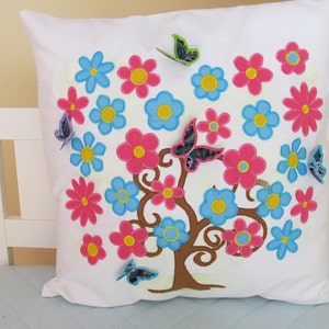 May include: White pillow with embroidered floral design. The design features a tree with pink, blue, and yellow flowers and butterflies.