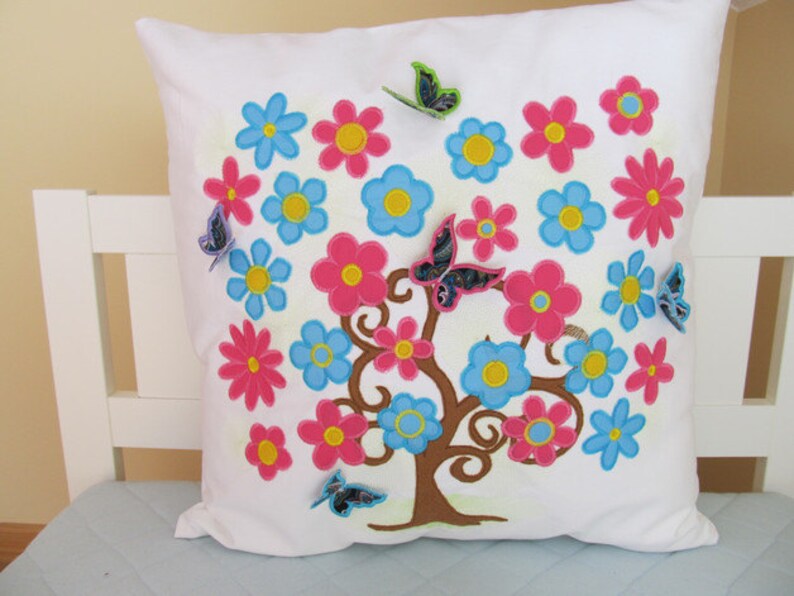 May include: White pillow with embroidered floral design. The design features a tree with pink, blue, and yellow flowers and butterflies.