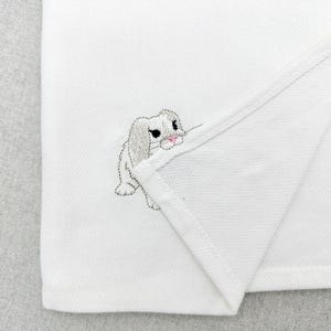 May include: White fabric with a subtle herringbone pattern, featuring an embroidered bunny in gray and pink. The bunny is positioned in the corner of the folded fabric, adding a touch of whimsy.