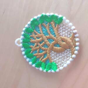 May include: A white, green, and gold embroidered ornament shaped like a circle with a tree design. The tree has a brown trunk and green leaves.