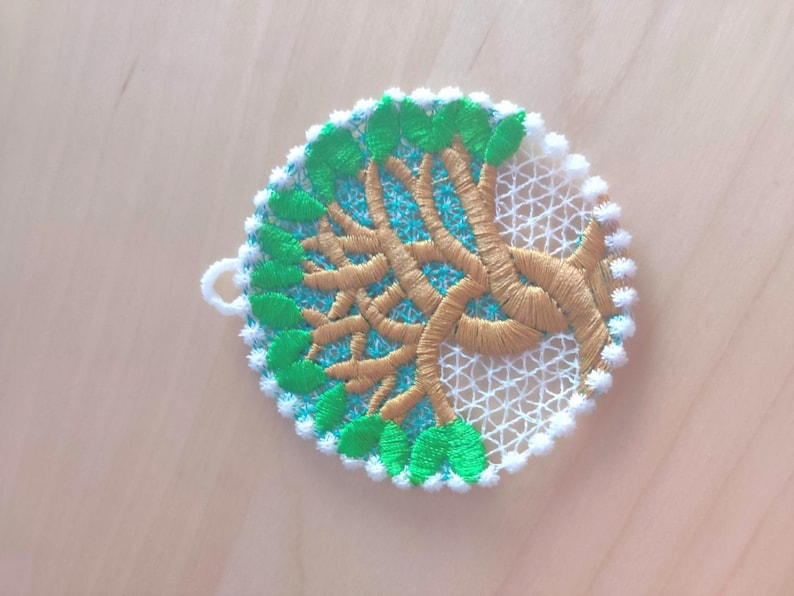 May include: A white, green, and gold embroidered ornament shaped like a circle with a tree design. The tree has a brown trunk and green leaves.
