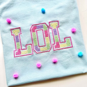 May include: A light blue t-shirt with the word "LOL" embroidered in colorful squares. The shirt is decorated with pink and blue pom-poms.