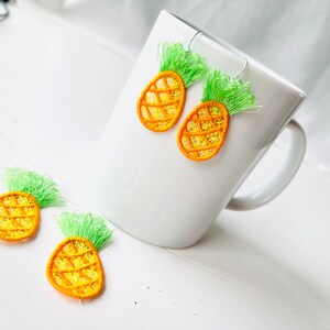 May include: Three pairs of pineapple-shaped earrings with green tops and yellow bodies. The earrings are made of embroidery thread and are hanging from a white mug.