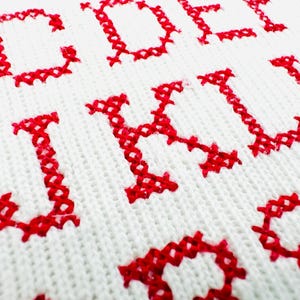 May include: Close-up of white fabric with red cross-stitch letters. The letters J, K, L, and M are visible, along with parts of other letters. The cross-stitch is detailed and the fabric has a textured appearance.