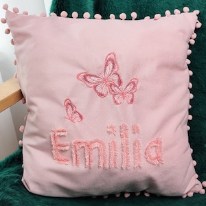 K&ouml;nnte beinhalten: Rosafarbenes Kissen mit Pompon-Besatz und gestickten Schmetterlingen sowie dem Namen "Emilia".